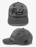 A black washed cotton baseball cap shown from the front and side angles against a light gray background. The front features the word 'Rebel' embroidered in black script, and the side displays a small Anarchy inspired embroidered 'R' logo.